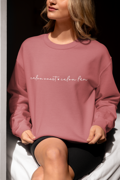 A woman smiling and wearing a dusty pink sweatshirt with the embroidered text 'calon onest é calon lan' in white.