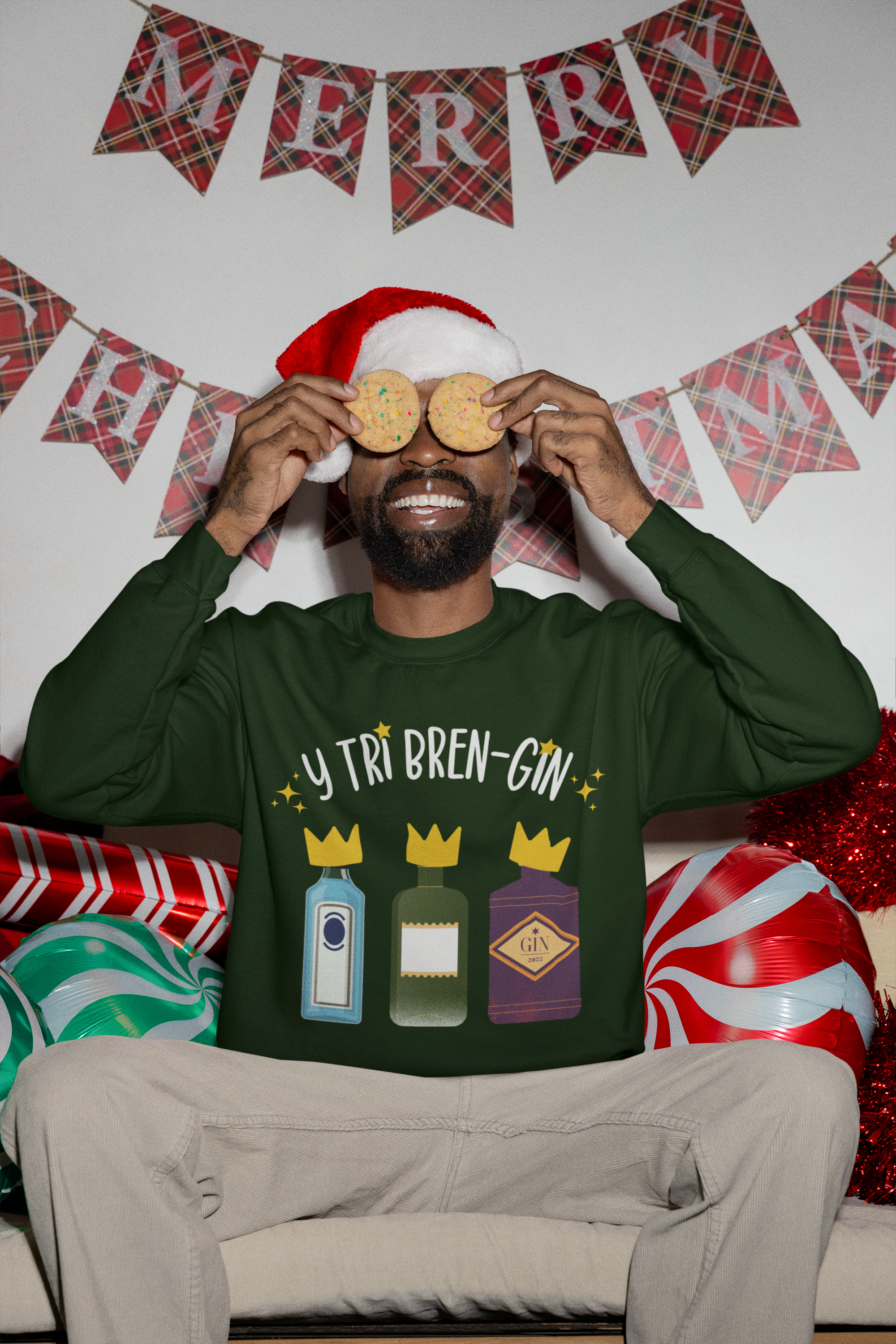 A person wearing a green Christmas sweatshirt with text and cartoon images of bottles, holding two cookie cutters shaped like glasses on their eyes, sitting in front of a decorated backdrop with festive decorations.