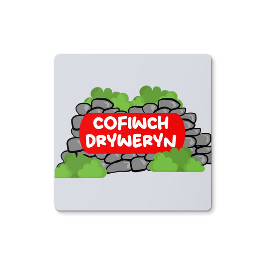 A square coaster with a green background featuring a red and white text that reads 'COFIWCH DRYWERYN' on a simulated stone wall design.